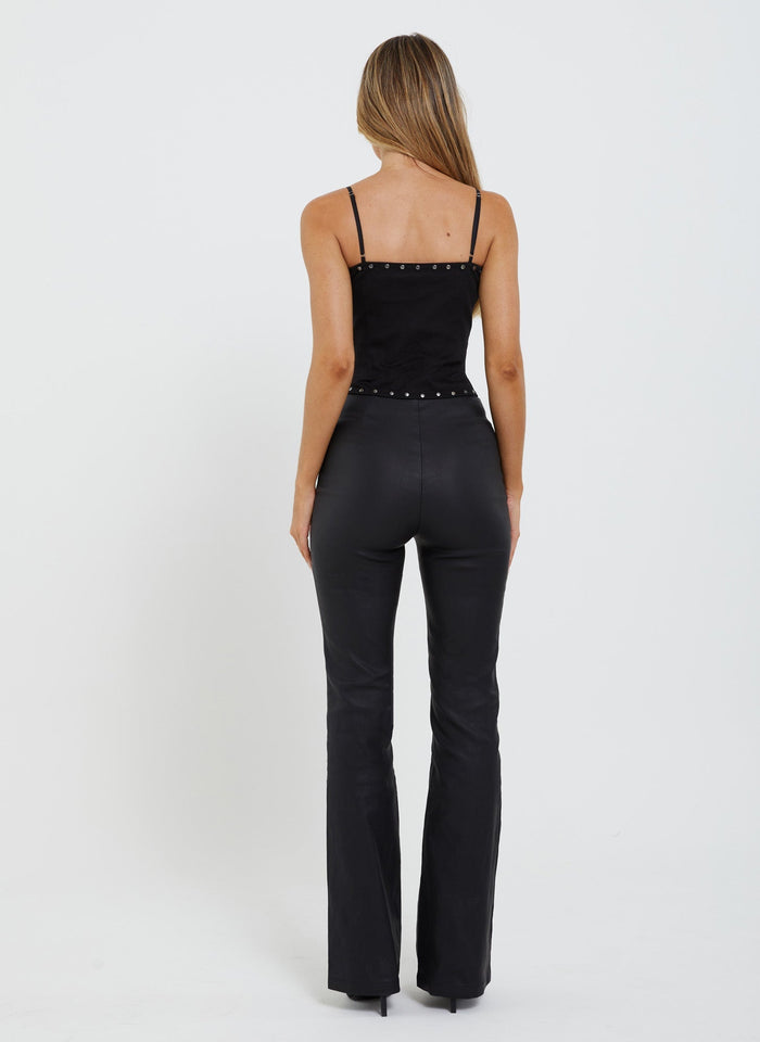 A woman with long, blonde hair stands with her back to the camera, donning a black, sleeveless top and the DOPHA PANT - BLACK by TIGERMIST. The outfit is accented with small studs on the top's neckline and waistline. The background is plain white.
