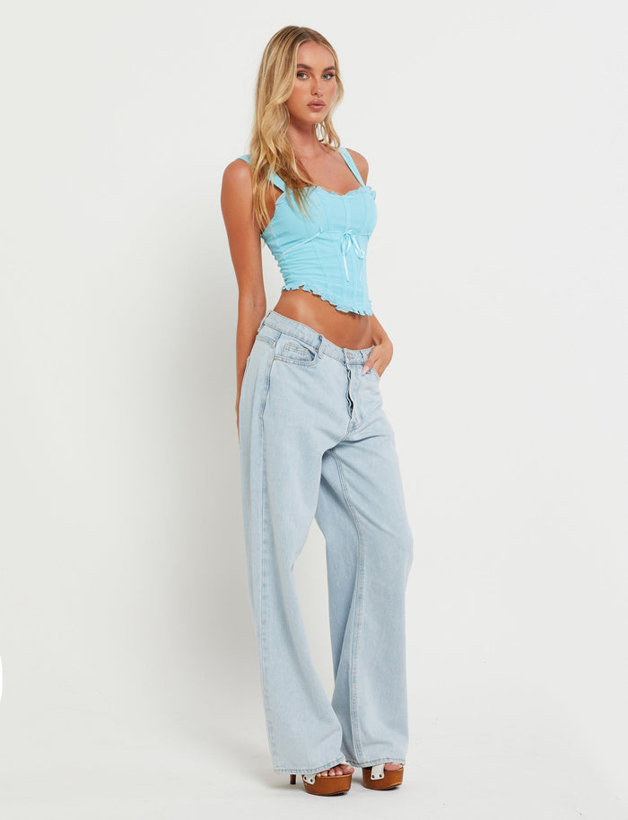 A woman poses against a plain white background, wearing the ELARA TOP in Baby Blue from TIGERMIST and high-waisted, light-wash, wide-leg Tiger Mist jeans. She has wavy blonde hair and is wearing white platform sandals. One hand is in her pocket while the other rests by her side.