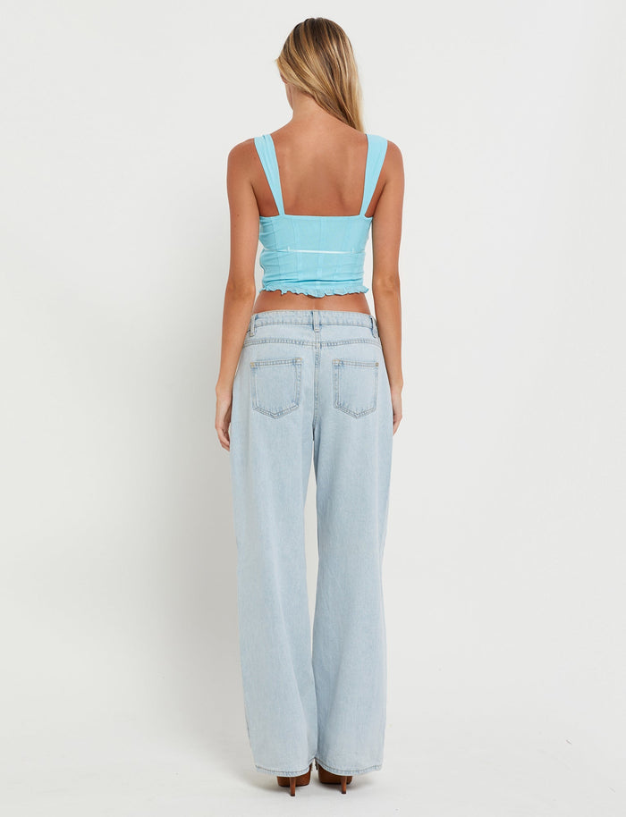 A person with long blonde hair is standing facing away, stylishly dressed in the ELARA TOP - BLUE (BABY BLUE) and high-waisted wide-leg jeans from TIGERMIST. The background is plain white.