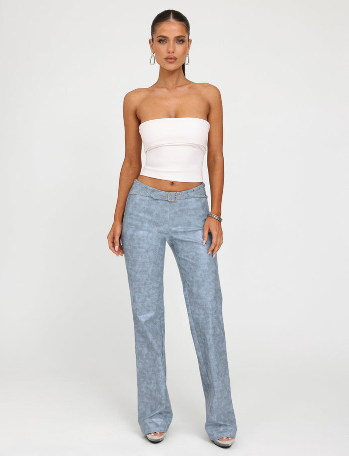 A person stands against a plain background, wearing a white strapless top and the TIGERMIST EMERSYN PANT in Dusty Blue, featuring a croc belt and flared silhouette. The individual has long hair tied back and accessorizes with hoop earrings, bracelets, and silver open-toe high heels. They are looking directly at the camera.