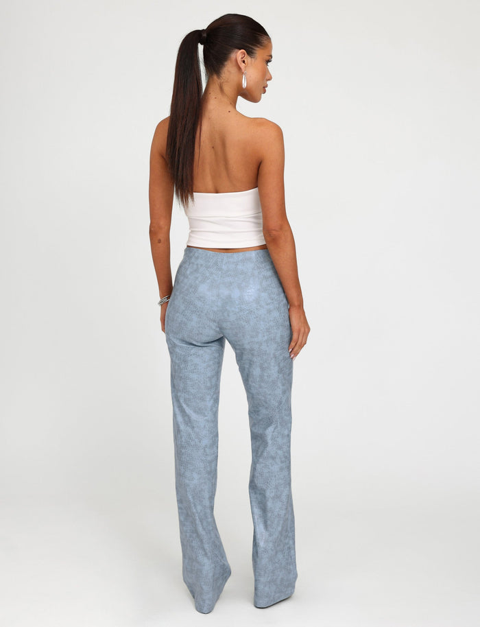 A woman is facing away from the camera, showcasing the back of her outfit. She is wearing a white tube top and high-waisted EMERSYN PANT in Dusty Blue from Tiger Mist. Her long, straight hair is tied back in a ponytail against a plain, light-colored backdrop.