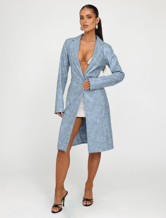 A woman stands against a plain, off-white background, wearing the TigerMist EMERSYN TRENCH COAT - BLUE : DUSTY BLUE with a deep V-neck over a white mini skirt. She accessorizes with dangly earrings and black high-heeled sandals. She gazes confidently at the camera with one hand on her coat.