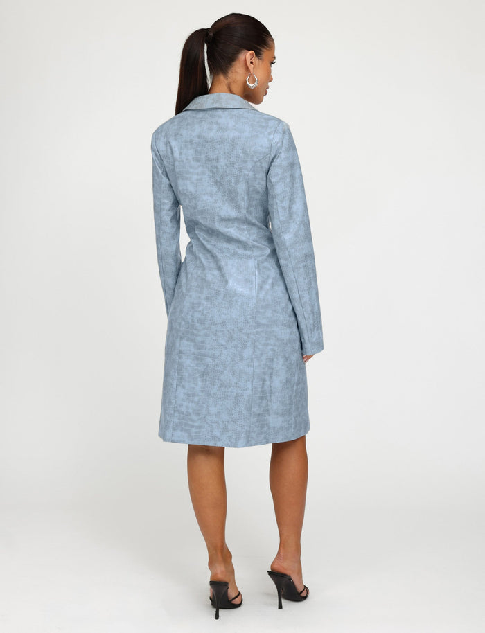 A woman with a ponytail is standing with her back to the camera, showcasing a TIGERMIST EMERSYN TRENCH COAT in dusty blue, paired elegantly with black high-heeled sandals against a plain white background.