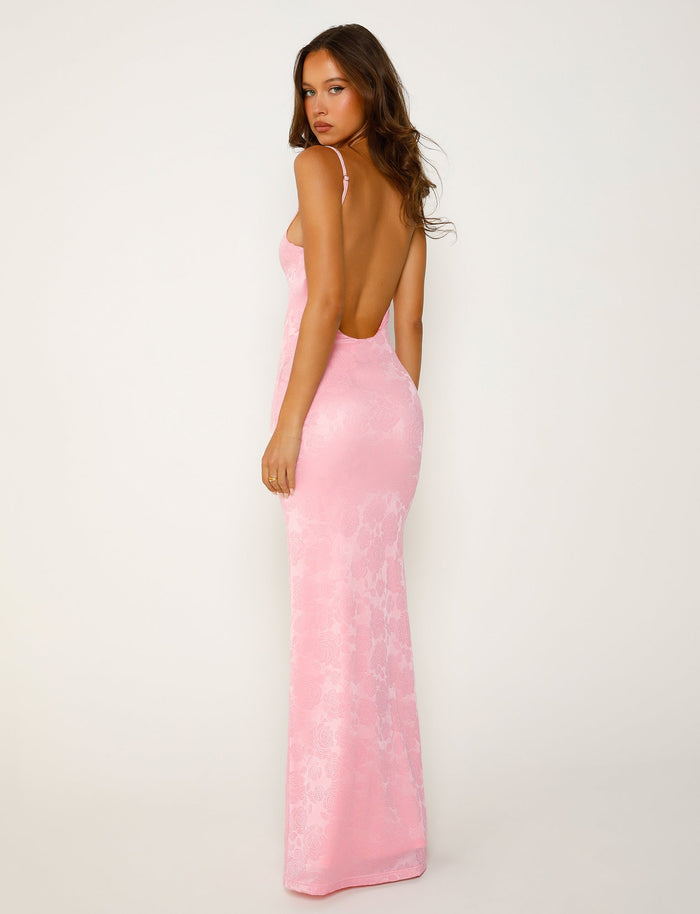 A woman in a form-fitting, backless ERICKA DRESS - PINK from TIGERMIST stands with her back turned to the camera. The dress features thin shoulder straps and flows down to the floor. She is gazing over her shoulder against a plain white background.