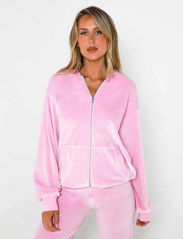 A woman is standing against a white background wearing the Baby Pink Ericka Hoodie by TIGERMIST, a velour tracksuit with a zipper. She has long blonde hair and her hands are in the pockets of her outfit, which features a relaxed fit with a slightly oversized jacket and pants, exuding casual elegance.