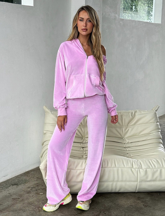 A person wearing an ERICKA HOODIE - PINK : BABY PINK and matching wide-leg pants from TIGERMIST stands indoors in front of a white leather sofa. They have long hair and are looking to the side with a neutral expression. The room has a minimalist design with light-colored walls.