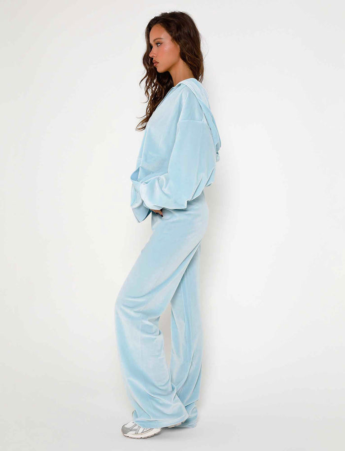 A woman with long, wavy hair stands in profile, facing left. She wears a light blue, hooded velour tracksuit from TigerMist featuring the ERICKA PANT in baby blue with wide-leg styling and holds her hands in her pockets. She completes the outfit with white sneakers. The background is a plain white wall.