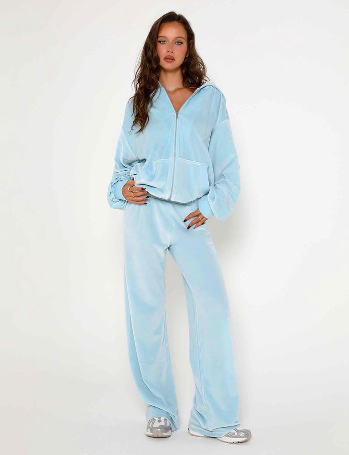 A person with long brown hair is standing against a plain white background, dressed in TIGERMIST's baby blue ERICKA PANTS with a matching velour hoodie and silver sneakers. One hand rests in a pocket of the hoodie, and they have a relaxed expression.