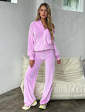 A woman with long blonde hair is standing indoors wearing the BABY PINK ERICKA PANT from TIGERMIST. She has one hand in her pocket and is leaning slightly against a cushioned beige chair, paired perfectly with white and neon yellow sneakers.