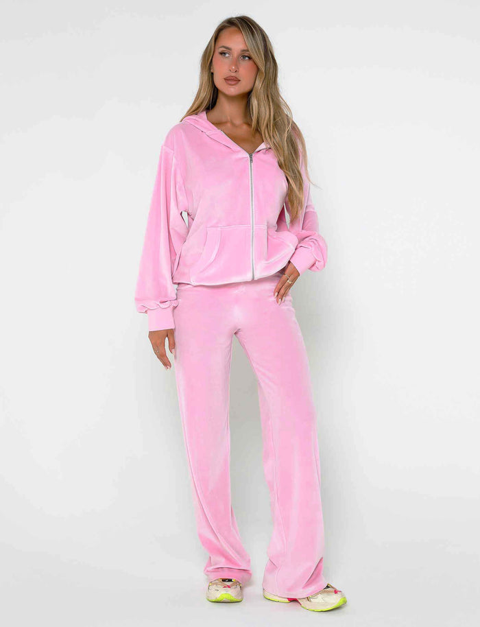 A person with long blonde hair is wearing a matching BABY PINK ERICKA PANT and zip-up hoodie from TIGERMIST. They are standing against a plain white background, one hand in their hoodie pocket, sporting white and yellow sneakers.