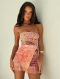 A woman with long brown hair wears the TIGERMIST ESME SKORT in a grunge floral print featuring pink and orange hues. She stands against a plain white wall, gazing directly at the camera. Silver bangles adorn her wrist, adding a touch of elegance to her bold look.