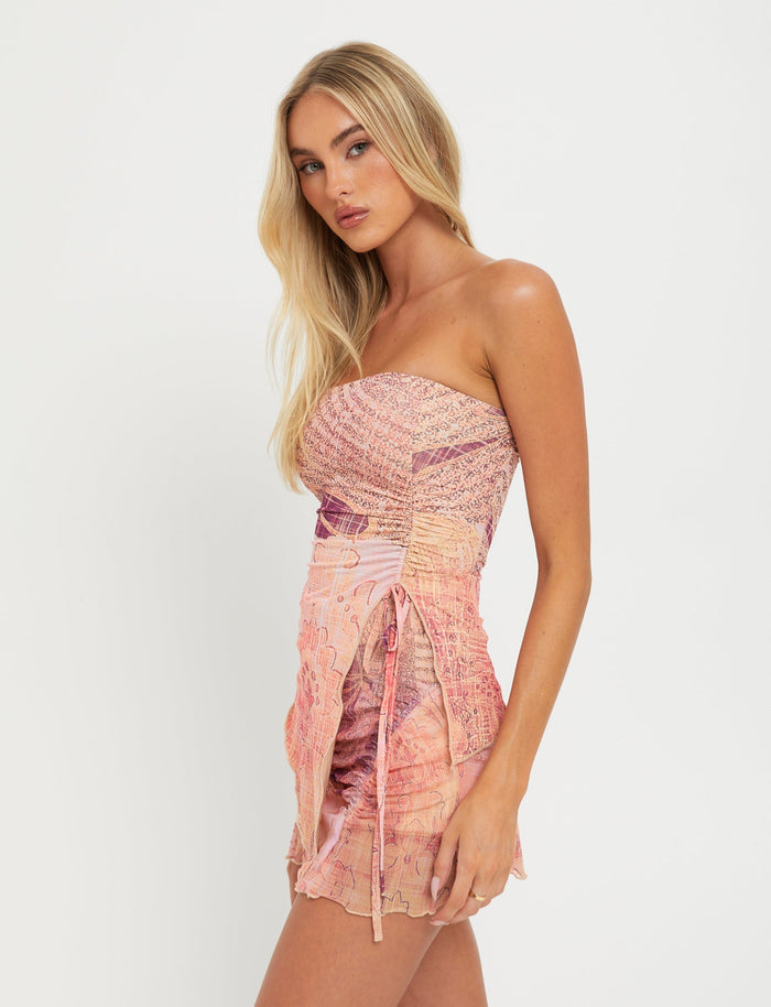 A woman with long blonde hair wears the TIGERMIST ESME SKORT in a grunge floral design, featuring a strapless pink and purple bodice that hugs her body in a bodycon fit. Set against a plain white background, the outfit boasts ruched detailing and a front tie, adding subtle touches of floral elegance.