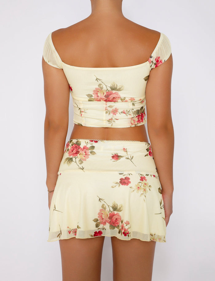 A person with their back to the camera is wearing a light yellow FEYRA TOP - YELLOW : MACRO FLORAL : POP FLORAL crop top and matching skirt from TIGERMIST. The outfit showcases a vibrant pattern of pink and red flowers with green stems and leaves. The individual has their arms relaxed at their sides against a white background.