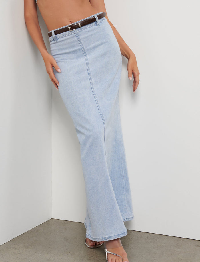 A person is leaning against a white wall, wearing a light blue, high-waisted FLETCHER SKIRT - BLUE by TIGERMIST with a brown belt and open-toe heeled sandals. Only the lower portion of their torso and legs are visible in the image.