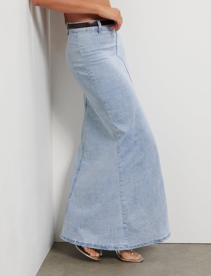 Side view of a person wearing the TIGERMIST FLETCHER SKIRT in denim blue, which is high-waisted with a flared bottom. The person is standing with one arm resting against a white wall and they are wearing clear, high-heeled shoes. The background is a plain indoor setting.