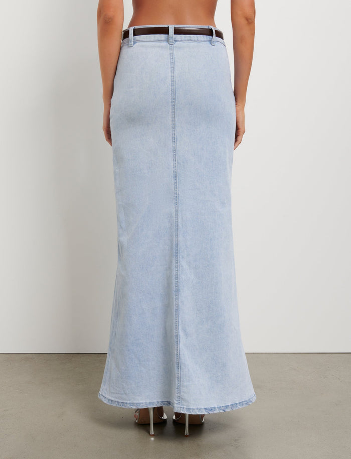 A person is standing with their back facing the camera, showcasing the TIGERMIST FLETCHER SKIRT - BLUE : DENIM BLUE, a high-waisted, light-wash denim maxi skirt with a back slit. Paired with a brown belt and heeled shoes, the ensemble captures TIGERMIST's signature style. The plain white background and visible concrete floor add to the minimalist aesthetic.