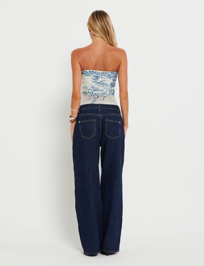 A woman with long blonde hair is shown from the back, wearing a strapless TIGERMIST HARRIETT TUBE TOP in blue with a tattoo print and high-waisted dark blue jeans. She has several bangles on her right wrist. The background is plain white.