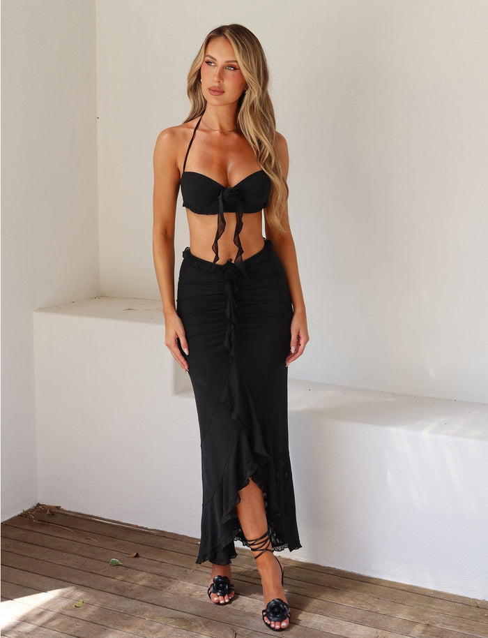 A woman with long blonde hair is standing against a white wall, wearing a stylish black bikini top and the HEAVEN MAXI SKIRT in black from TIGERMIST that features a ruffled hem. She pairs it with black sandals and looks towards the camera. A small leaf is visible on the wooden floor.