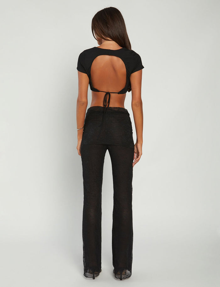 A woman stands facing away, showcasing her TIGERMIST outfit. She is wearing a black crop top with a circular cutout on the back and the INAYAH PANT - BLACK from Tiger Mist. Her straight, long hair flows down her back, and her arms are at her sides. The background is plain and light-colored.