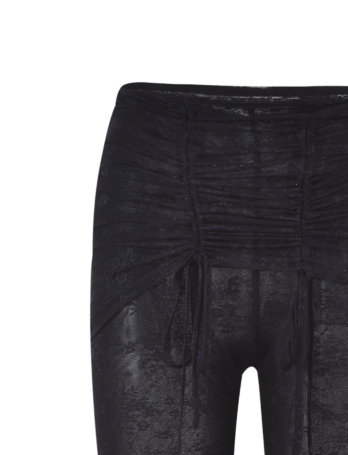 A close-up image of the INAYAH PANT - BLACK from TIGERMIST, featuring an elasticated waistband and ruching details near the waist. The fabric boasts a sheer, lacy texture, complemented by decorative drawstrings on both sides for an adjustable fit.