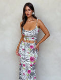 A woman with long, dark hair is wearing the IRIE TOP - MULTI : ZEBRA from TIGERMIST. The top features a fitted, animal print design with pink roses, thin spaghetti straps, and midriff cutouts. She is standing in front of a beige textured wall and looking directly at the camera.