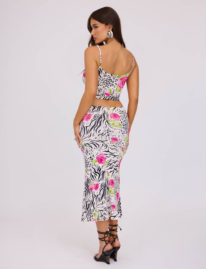 A woman is modeling the TIGERMIST IRIE TOP - MULTI : ZEBRA, a two-piece outfit featuring a crop top and high-waisted skirt adorned with a mixed pattern of zebra stripes, leopard spots, pink roses, and green leaves. She completes her look with black strappy heels and hoop earrings while posing in a white studio setting.