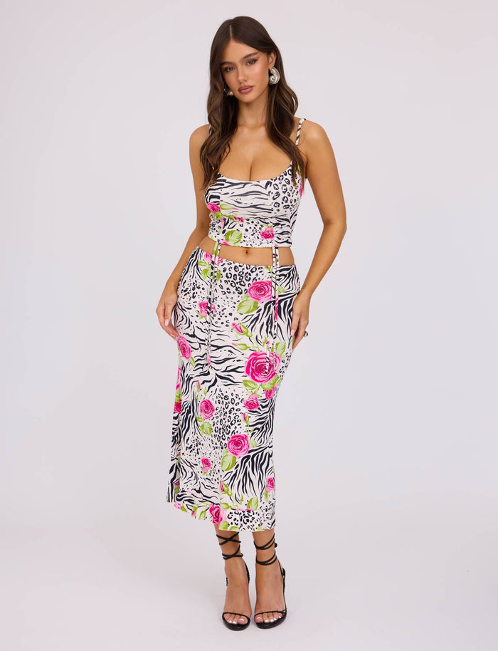 A woman stands in a white studio wearing the mid-length, form-fitting IRIE TOP - MULTI : ZEBRA from TIGERMIST. The top features a striking mix of zebra print and floral patterns in black, white, and vibrant pink. It has spaghetti straps and cut-out sides. She has long, wavy hair and complements her outfit with black strappy high heels.