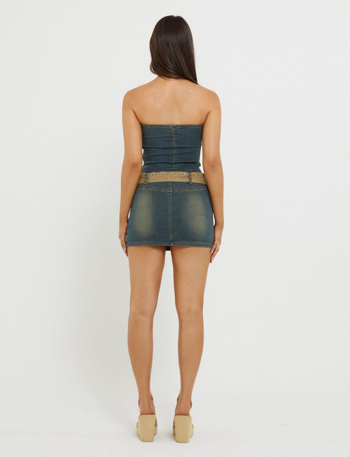 A person with long brown hair stands facing away from the camera in a fitted TigerMist ISABEL SKORT - BLUE, ending above the knees, paired with a matching belt with metal eyelets and tan platform shoes. The background is plain white.