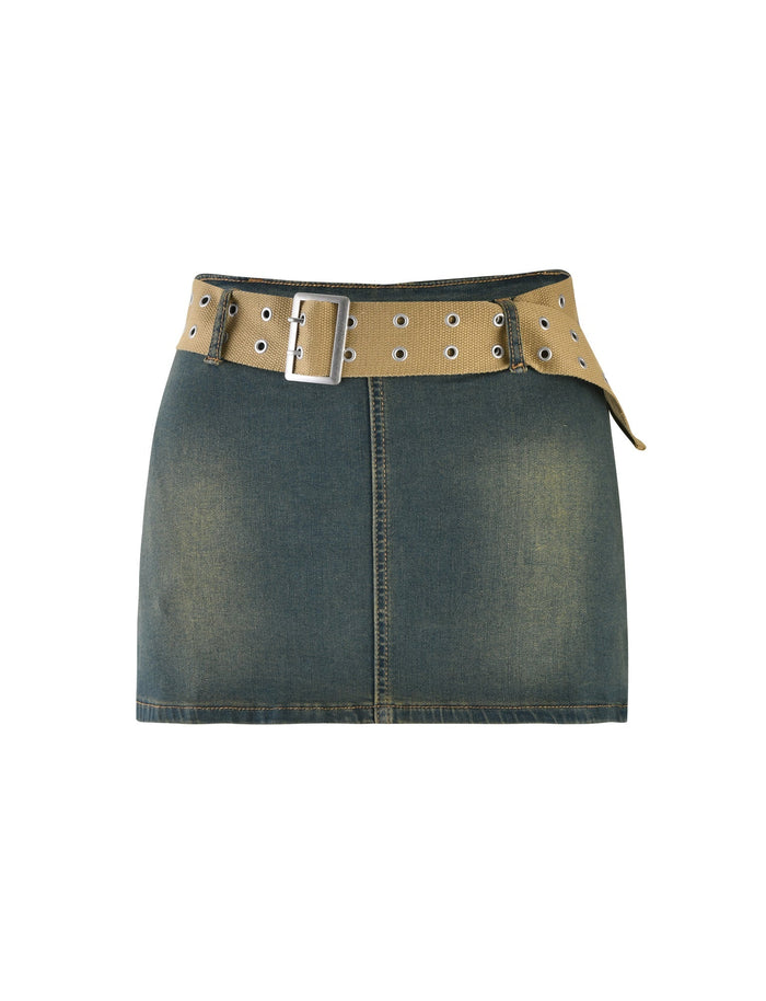 The ISABEL SKORT - BLUE by TIGERMIST is a high-waisted, dark denim mini skirt with a wide, light brown, grommeted belt featuring a silver buckle. The skirt has a slightly faded, worn look. The belt is looped through the waistband and hangs slightly on the right side.