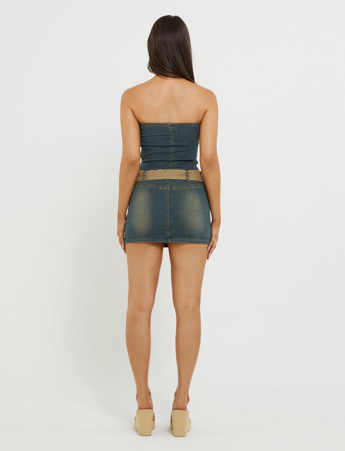 A woman with long, dark hair stands facing away from the camera. She is wearing the ISABEL TUBE TOP - BLUE : DENIM BLUE by TIGERMIST, paired with a wide, studded belt and beige wedge shoes. The background is white and plain.