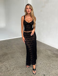 A woman with long blonde hair is wearing the ISADORA SKIRT - BLACK : LACE and a coordinating black lace crop top from TIGERMIST. She stands indoors on a concrete floor, with a neutral grey wall in the background. She gazes at the camera, one hand resting on her skirt and the other by her side.