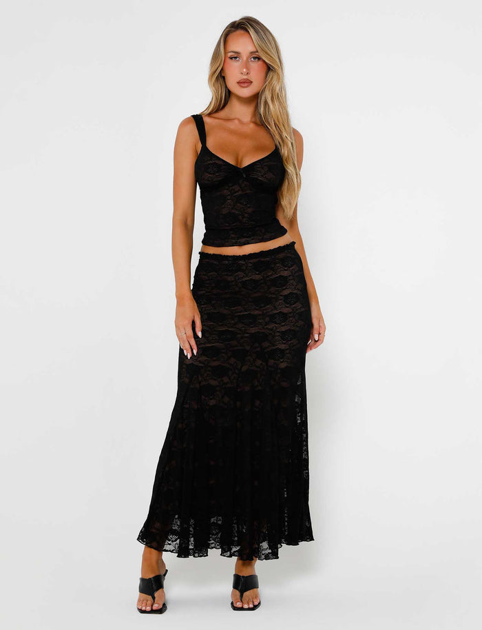 A woman with long, blonde hair, wearing TigerMist's stylish ISADORA SKIRT - BLACK lace skirt and a matching sleeveless top, poses against a plain white background. She is also sporting black high-heeled sandals. Her outfit features intricate floral patterns.