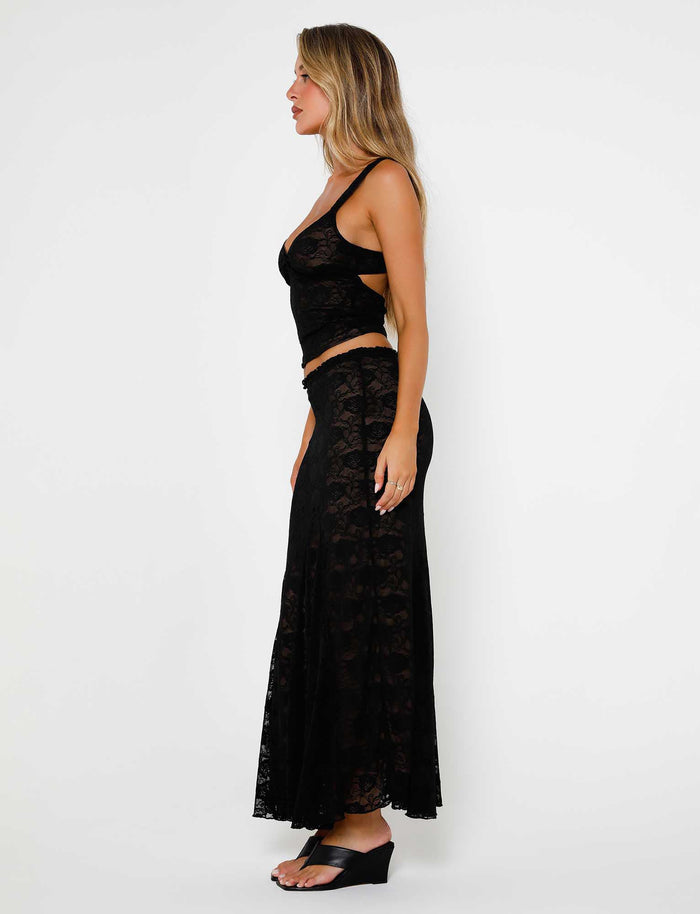 A woman with long hair is wearing the ISADORA SKIRT - BLACK : LACE and a matching black, sleeveless lace top. She is standing sideways, facing to the left, and wearing black wedge sandals by TIGERMIST. The background is plain white.