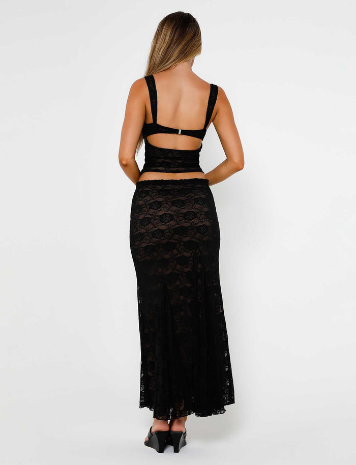 A woman with long brown hair is facing away from the camera, wearing a stylish ensemble from TIGERMIST: a black backless top paired with the ISADORA SKIRT - BLACK : LACE. She completes the look with elegant black heeled shoes. The background is plain white.
