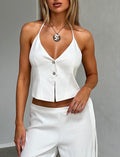 A woman is wearing the chic JAYLIN VEST - WHITE from TIGERMIST, featuring a halter-neck design with thin straps and two front buttons, paired with white pants. She accessorizes with a silver pendant necklace and stands in front of a plain light gray backdrop, her right hand resting on her hip.