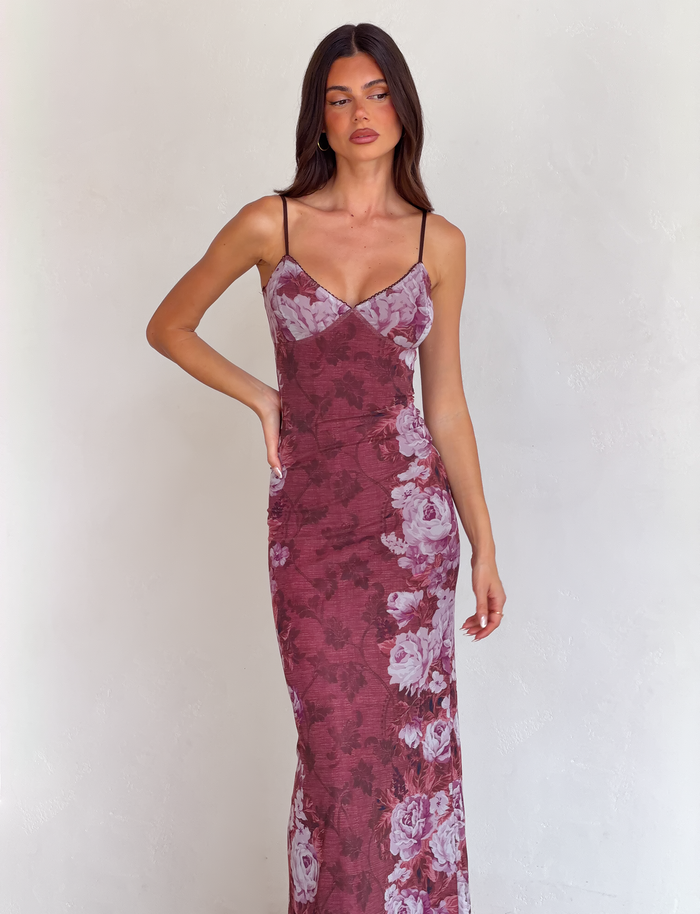 A woman in the TIGERMIST JULIANNA DRESS - MULTI: FLORAL: ROSE STRIPE, a sleeveless dress with spaghetti straps and a rose stripe floral print, stands against a light textured wall, hand on hip and gazing aside.