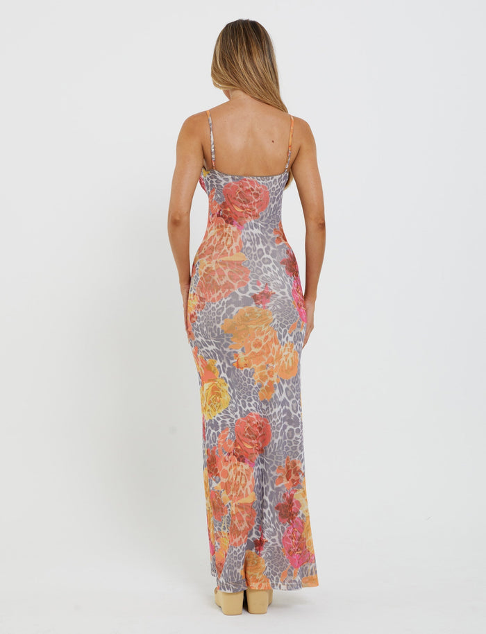 A person with long, light-brown hair is standing with their back to the camera, wearing a sleeveless, form-fitting TIGERMIST JULIANNA DRESS in a multi-colored floral and orange leopard print pattern. The dress is full-length and paired with beige high heels.
