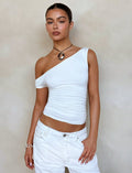 A person poses against a beige background, wearing the KADENCE TOP - WHITE and matching white pants from TIGERMIST. They accessorize with a simple black choker necklace featuring a pendant and a silver bracelet on their left wrist. Their hair is pulled back, highlighting their confident expression.