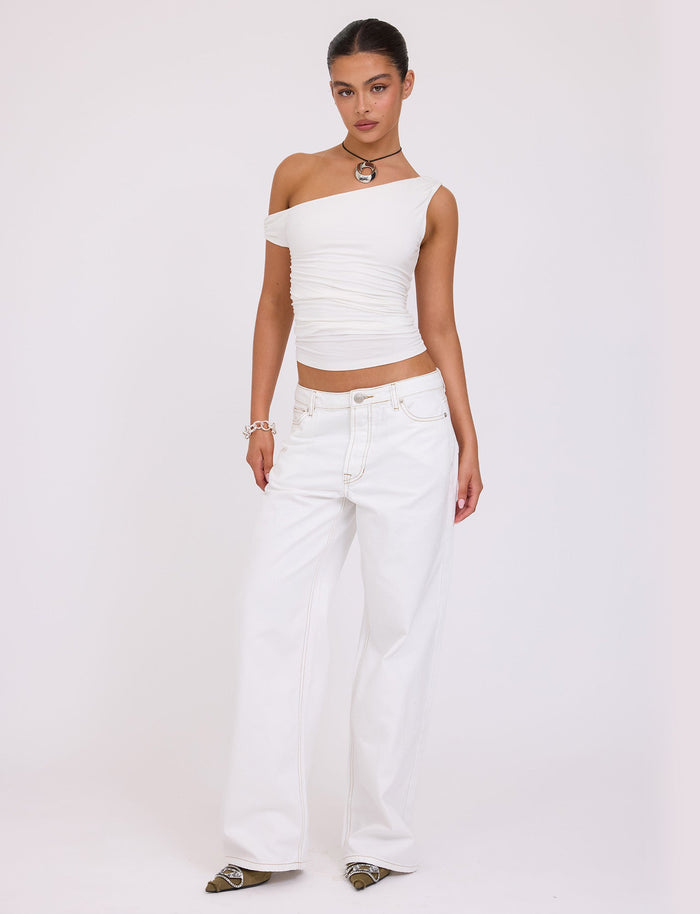 A person with their hair tied back stands against a plain background, donning a chic TIGERMIST KADENCE TOP - WHITE and loose, high-waisted white jeans. They accessorize with a necklace, bracelet, and pointed shoes, striking a relaxed pose.