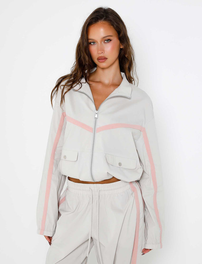 A woman is posing against a white backdrop, wearing the Dove Grey KAHANNA JACKET from TIGERMIST. The light beige tracksuit features pink accents and consists of a zip-up jacket with front pockets, along with matching drawstring pants. She has long, wavy brown hair and is looking directly at the camera.