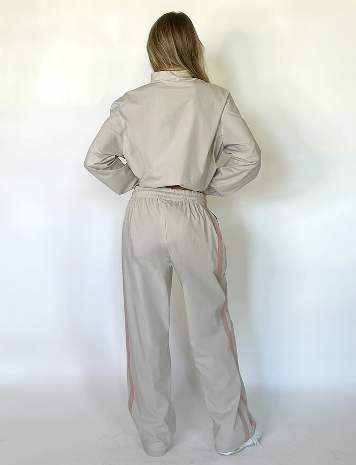 A person with long hair stands with their back to the camera, dressed in a chic TIGERMIST KAHANNA PANT in Dove Grey, featuring stylish pink stripes down the sides of the sleeves and pants. The background is plain and light-colored.