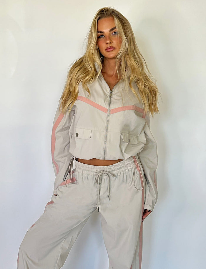 A woman with long, wavy blonde hair is wearing a TigerMist light-colored tracksuit with pink accents. The tracksuit includes a zip-up jacket with front pockets and the matching KAHANNA PANT in DOVE GREY. She poses against a plain white background, looking into the camera.