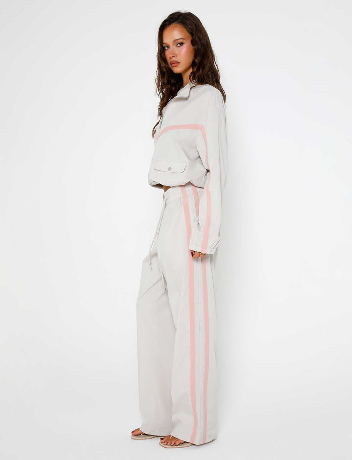 A woman with long, wavy hair is standing against a plain white background. She is wearing a matching light-colored long-sleeve top and the KAHANNA PANT - GREY : DOVE GREY from TIGERMIST, both adorned with pink stripes down the sides. She is looking to her left with a neutral expression.