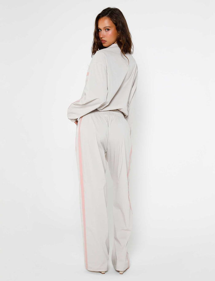 A person with long wavy hair is standing and looking over their right shoulder, dressed in a light gray outfit from TIGERMIST. They are sporting the KAHANNA PANT in Dove Grey, featuring a loose long-sleeve top and wide-legged pants adorned with subtle pink stripes down the sides. The plain white background keeps the focus on their stylish ensemble.