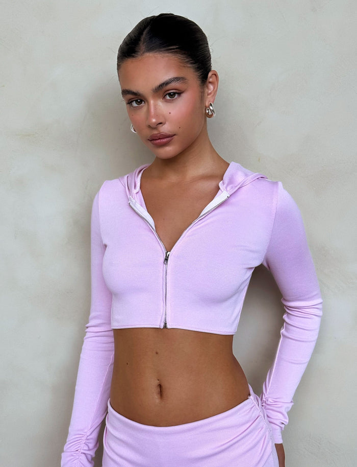 A woman with dark hair pulled back is wearing the KARINA JUMPER - PINK from TIGERMIST. She is standing against a plain, beige background and looking at the camera with a neutral expression. She accessorizes with small hoop earrings.