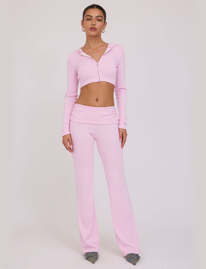 A woman stands against a plain white background wearing a light pink, long-sleeve crop top with a hood by TIGERMIST, paired with the matching KARINA PANT - PINK : BLUSH and grey shoes. She has her hair tied back and is facing forward with a neutral expression.