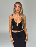 A woman with long blonde hair stands against a plain background, wearing the TIGERMIST KATINA CAMI - BLACK, featuring thin straps and a matching skirt. The cami showcases a cutout detail in the front and floral accents. She has a neutral expression, with her hands resting by her sides.