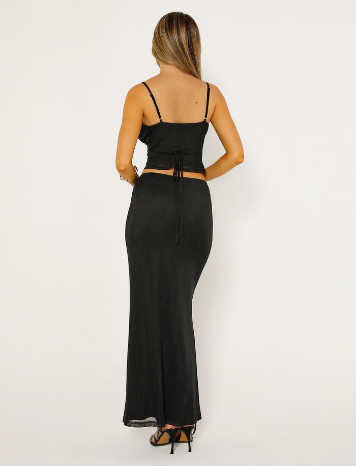 A woman, with long straight hair, stands facing away from the camera. She is dressed in a TIGERMIST black sleeveless top paired with the KATINA SKIRT - BLACK by TIGERMIST and wears black high-heeled sandals. The background is a plain white backdrop.