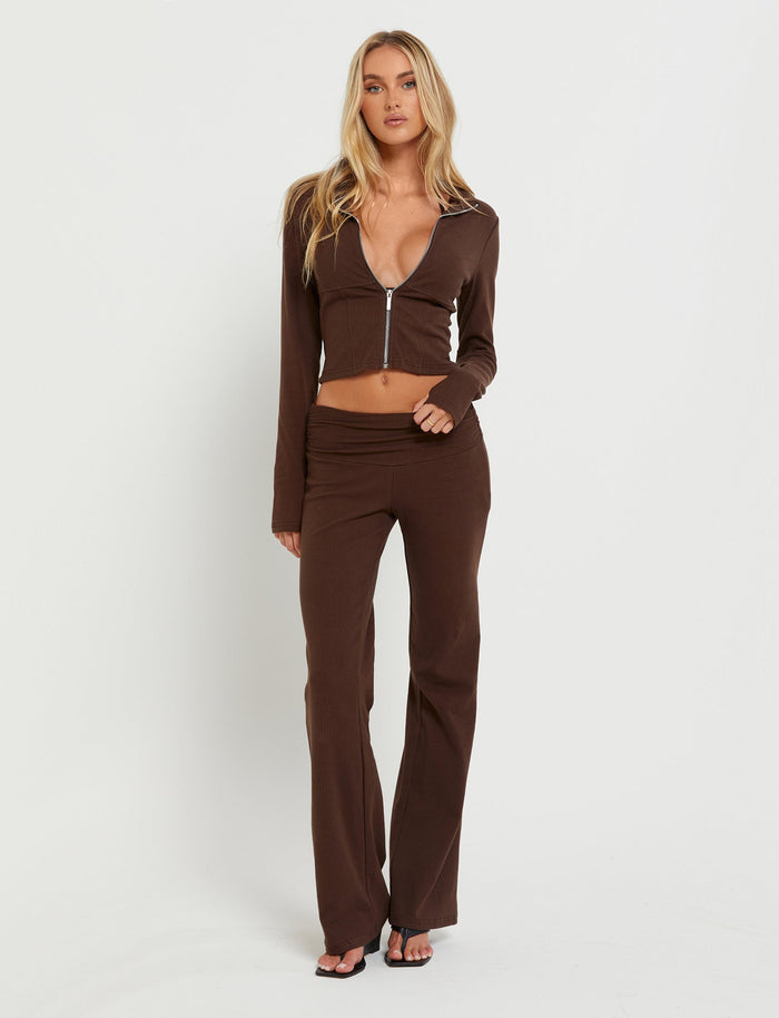 A woman with long blonde hair is posing against a plain white background, showcasing a chic TIGERMIST ensemble. She's wearing the Kavat Jacket in brown, complemented by matching high-waisted flared pants and accessorized with black sandals to complete her stylish look.