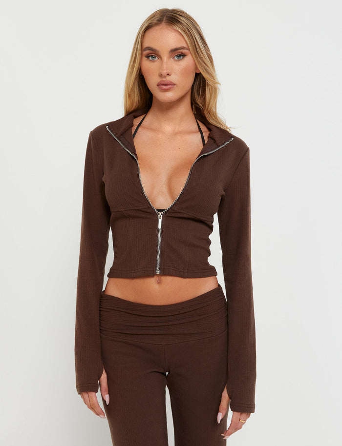 A woman with long blonde hair is wearing the KAVAT JACKET - BROWN from TIGERMIST, partially zipped to reveal a black undergarment, along with matching brown bottoms. She is posing against a white background, looking directly at the camera with a neutral expression.